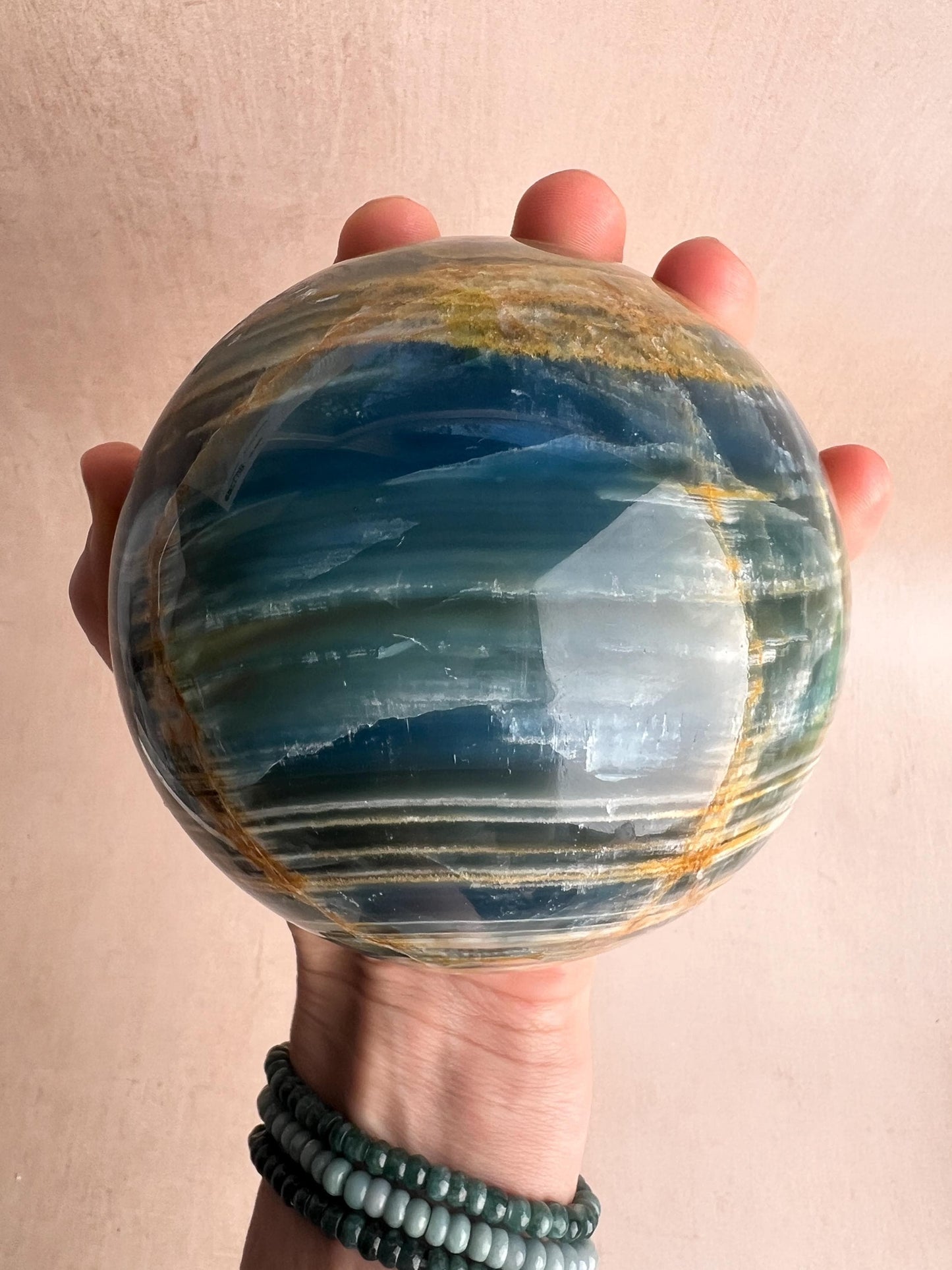 May include: A polished, spherical gemstone with bands of blue, green, and gold. The orb is held in a hand, showcasing its smooth, reflective surface. The gemstone is a decorative item.